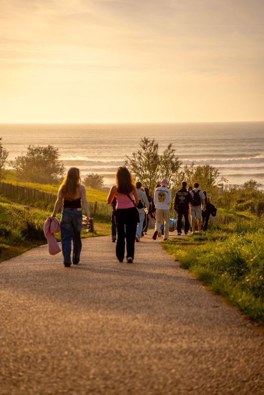Ride It — groupe descendant un chemin vers l'océan au coucher de soleil, planches à la main