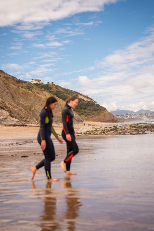 Ride It — deux rideuses en néoprène sur la plage à marée basse, falaise basque au fond